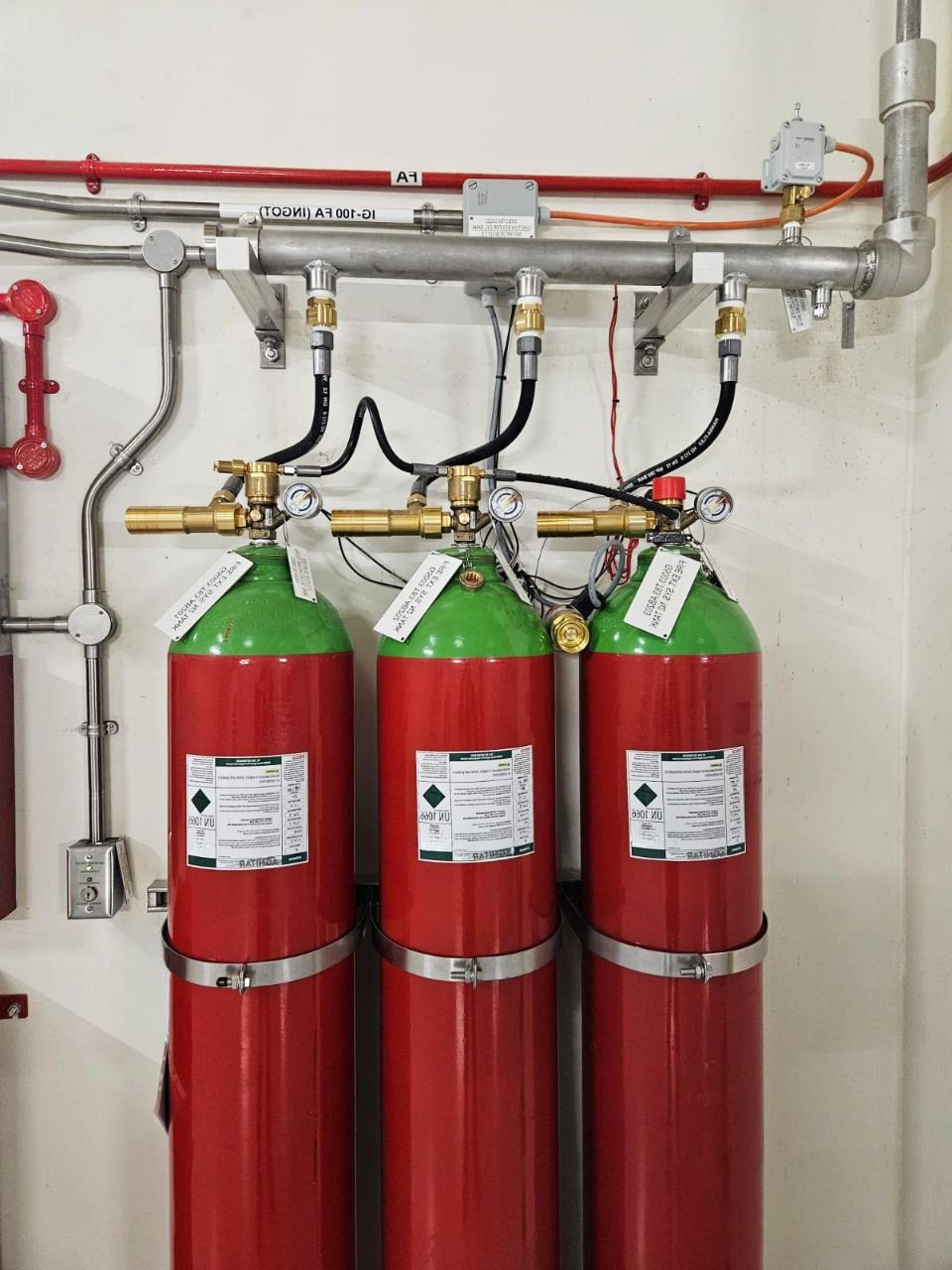 Three red fire suppression cylinders with green tops, connected to a silver metal pipe system with brass valves and black hoses, mounted on a white wall.
